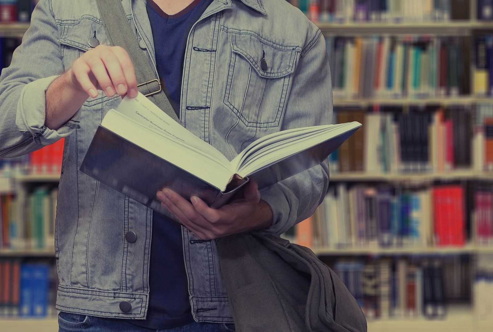 Detail of a student in a denim jacket leafing through a large reference book in a library